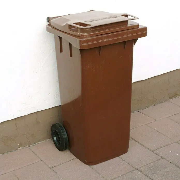 Floraworld Mülltonne mit Deckel Cargo - 120 l, Braun - Braune Mülltonne aus Kunststoff mit schwarzem Rad, die an einer weißen Wand steht, zur Entsorgung von Abfällen im Außenbereich.
