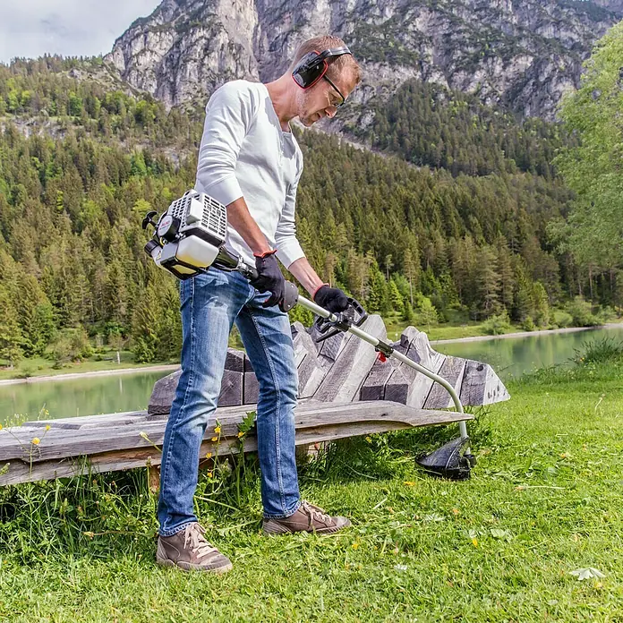 AL-KO Benzin-Motorsense BC 223 L-S - 700 W, Schnittbreite Faden: 41 cm - Ein Mann mit einer Motorsense trimmt Gras in der Nähe einer Bank mit einer Bergkulisse im Hintergrund.