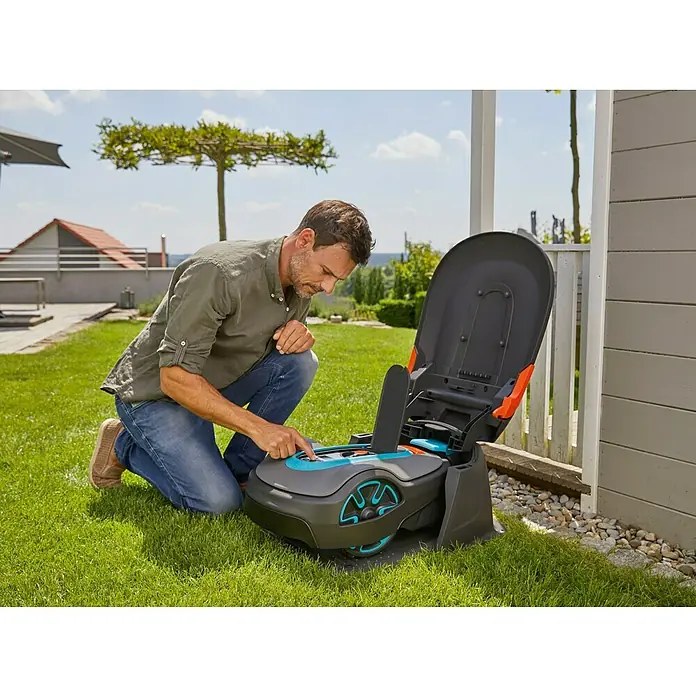 Gardena Mähroboter-Garage Sileno Life - L x B x H: 790 x 590 x 333 mm, Grau, Gardena Mähroboter Sileno minimo/Sileno life/Sileno city/smart Sileno life/smart Sileno city - Mann bedient einen Gardena Mähroboter auf einer grünen Rasenfläche in der Nähe eines Hauses mit weißer Fassade.