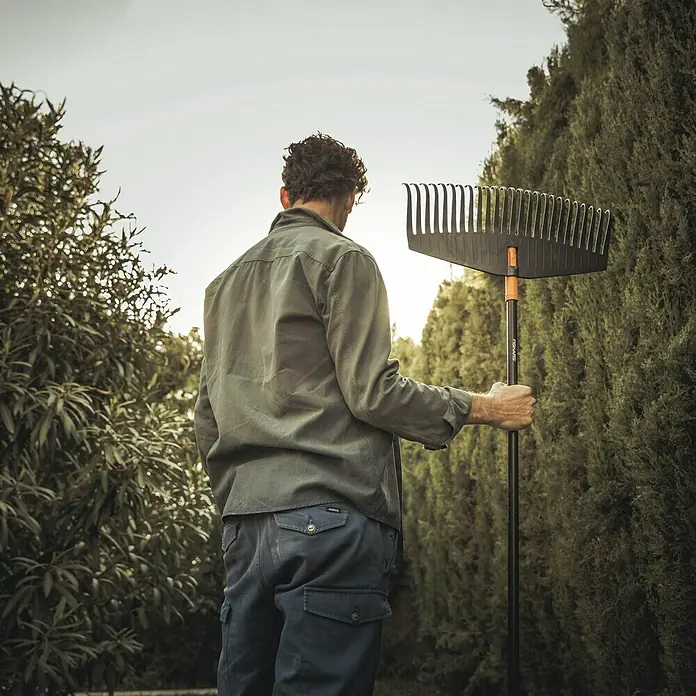 Fiskars Solid Laubbesen - Arbeitsbreite: 52 cm, Anzahl Zinken: 24, Art Stiel: Langstiel - Ein Mann hält einen Fiskars Rechen vor einem Garten. Der Rechen ist schwarz mit einem orangefarbenen Griff.