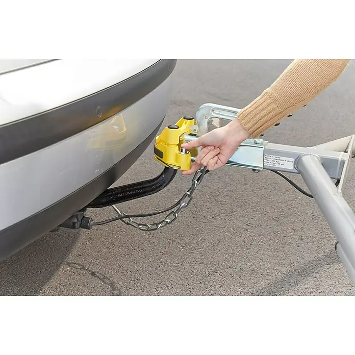Mottez Dispositivo antirrobo A075B - L x An x Al: 20 x 9,5 x 10 cm - Imagen de una persona que conecta un bloqueo de enganche de remolque amarillo a un coche, ofreciendo seguridad.