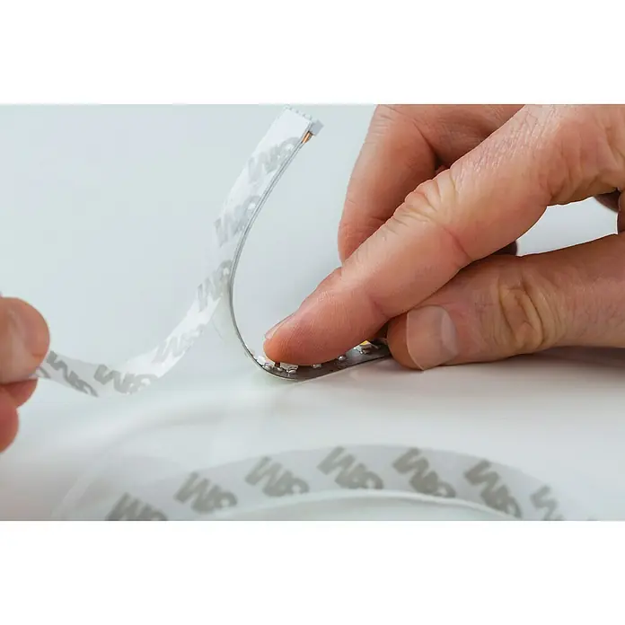 Paulmann MaxLED 1.000 LED-Band Basisset unbeschichtet - Länge: 3 m, Lichtfarbe: Warmweiß, 40 W, 3.300 lm - Nahaufnahme einer Person, die die Schutzfolie von einem LED-Streifen mit 3M-Kleber entfernt, um ihn zu befestigen.