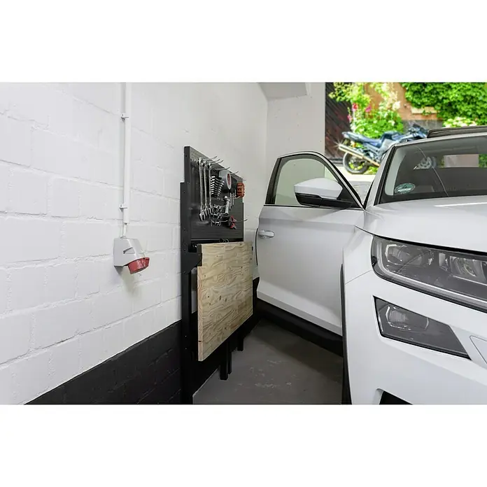 Werkbank Klappbar - L x B: 64,6 x 121,5 cm, Arbeitshöhe: 142 cm, Belastbarkeit: 150 kg - Nahaufnahme eines Garagenarbeitsplatzes mit Werkzeugwand und Werkbank neben einem weißen Auto. Praktische Raumlösung.
