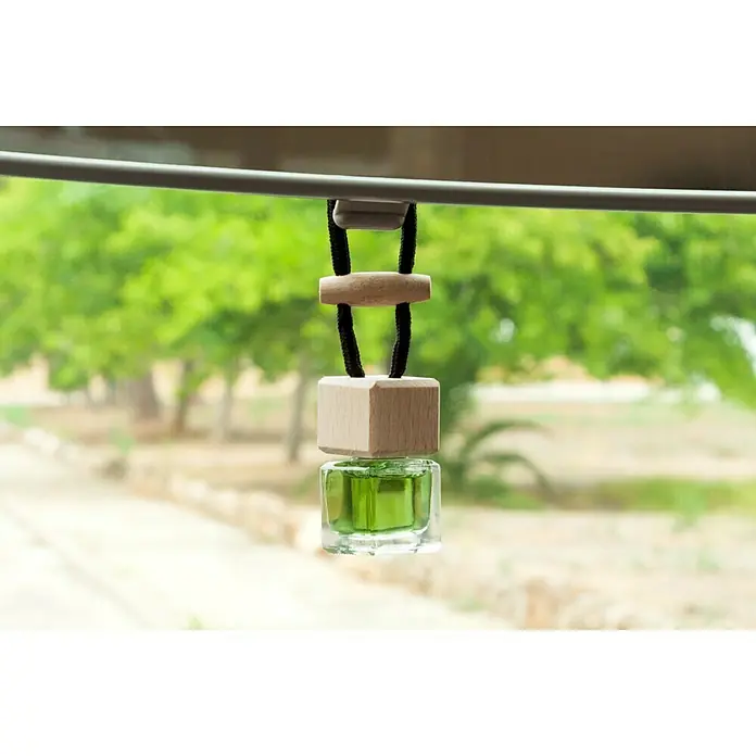 Ambientador de coche para colgar - Manzana, 8 sem. - Un ambientador cuadrado con detalles de madera y líquido verde cuelga contra un paisaje verde y borroso.