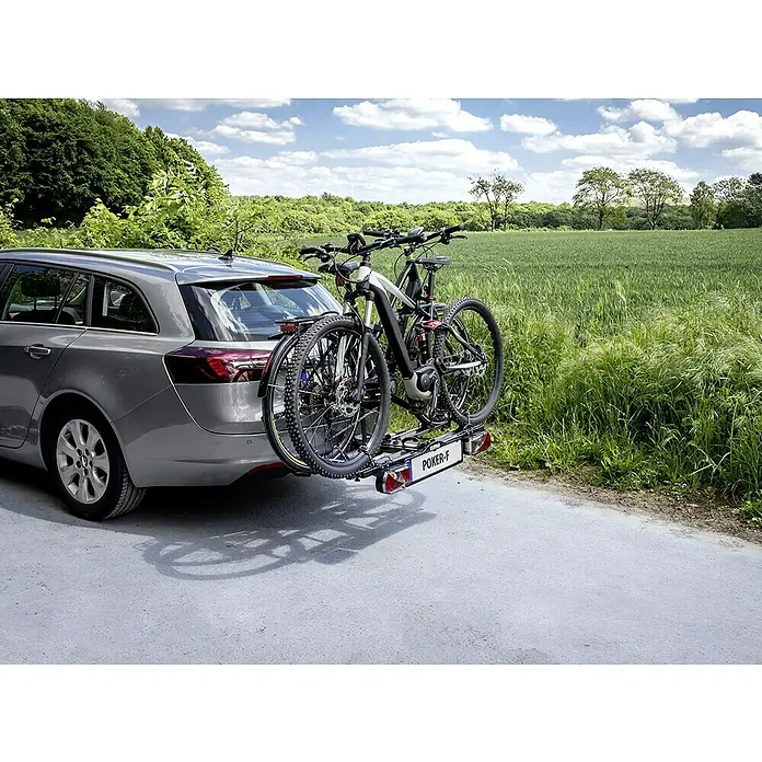 Eufab Fahrradträger Poker-F - Geeignet für: 2 Fahrräder, Traglast: 60 kg, Passend für: Fahrzeuge mit Anhängerkupplung - Ein grauer Kombi transportiert zwei Fahrräder auf einem Heckträger der Marke "POKER-F", während er auf einer Straße in ländlicher Umgebung parkt.