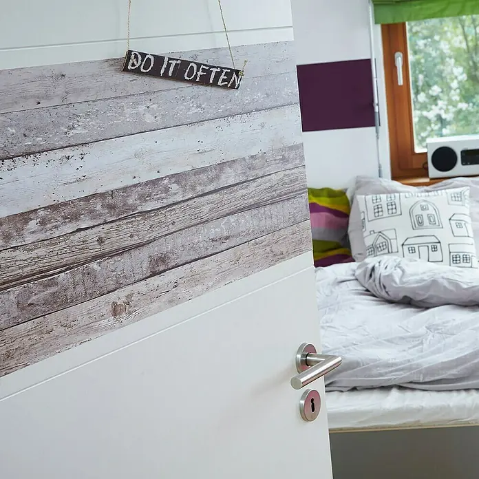 A.S. Création Designpanel pop.up - Bretter, Beige/Braun, 35 cm x 2,5 m - Nahaufnahme einer Zimmertür mit „Do It Often“-Schild, das an der Tür hängt, und Einblick in ein Schlafzimmer mit Bett.