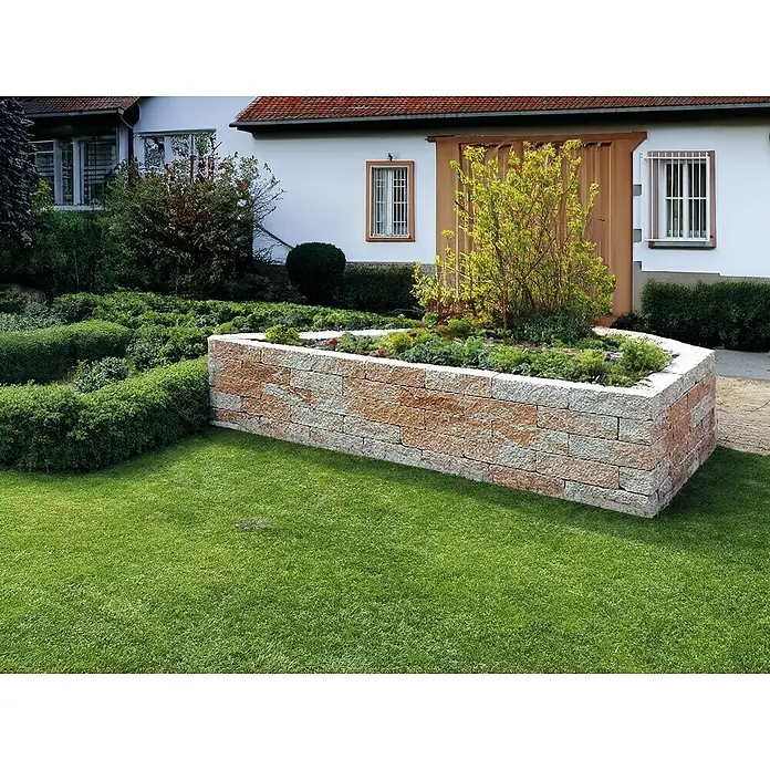 EHL Mauerstein Anzio Light - 50 x 18 x 15 cm, Sandstein, Beton - Ein erhöhter Pflanzkasten aus Natursteinblöcken mit üppiger Bepflanzung, eingebettet in einen gepflegten Garten vor einem charmanten Haus.