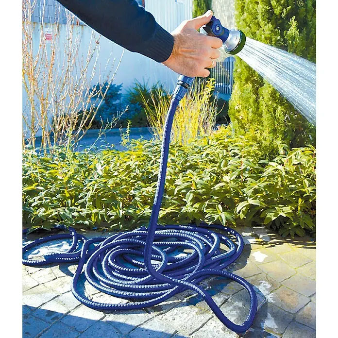 Manguera extensible con pistola 8F - Largo: 7,5 m - Manguera de jardín en uso: Un hombre riega plantas con una manguera de jardín azul flexible y una boquilla de rociado, que produce un fuerte chorro de agua.