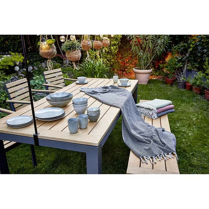 Sunfun Gartentisch Lynn - L x B x H: 220 x 100 x 76 cm, Akazie, Aluminium, Teakbraun, Anthrazit - Ein Gartentisch mit Geschirr und einer Decke, die elegant drapiert ist. Hängende Pflanzen und Lichter schmücken den Hintergrund.