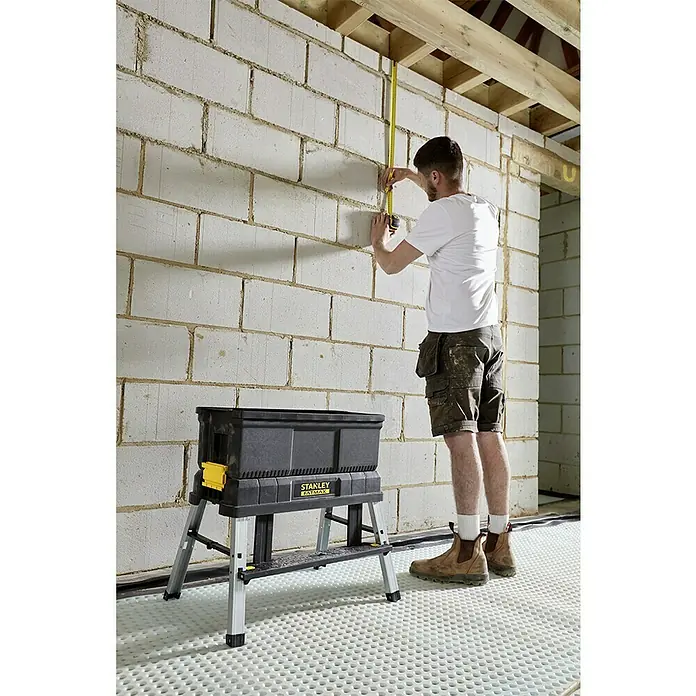 Stanley FatMax Werkzeugkasten FMST81083-1 - L x B x H: 29,6 x 64 x 28,7 cm, Klappbar - Ein Mann misst mit einem gelben Bandmaß die Wand, während ein Stanley Fatmax Werkzeugkasten-Arbeitstisch vor ihm steht.