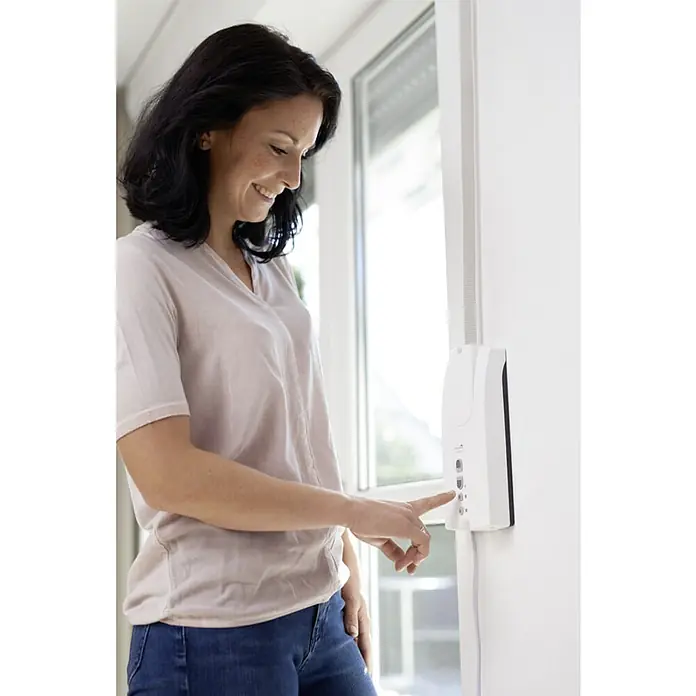 Schellenberg Recogedor de cinta de persiana Rollodrive 55 - En pared, Anchura de la correa: 14 mm - 23 mm, 230 mm - Una mujer opera un dispositivo electrónico blanco en una pared. Es un control automatizado de persianas, simplificando la vida.