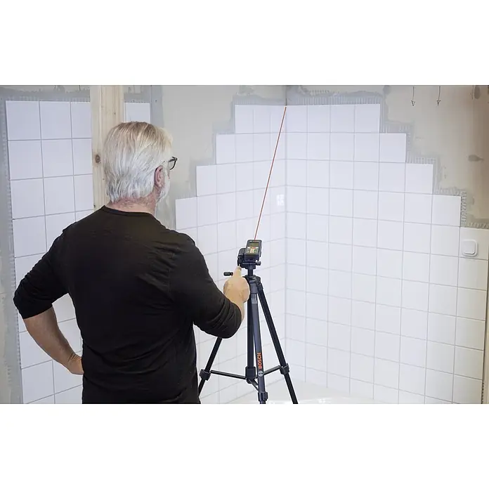 Bosch Professional Laserentfernungsmesser GLM 80 + Stativ BT150 - Messbereich: 0,05 - 80 m, Mit Stativ - Ein Mann justiert einen Bosch Linienlaser auf einem Stativ, um Fliesenarbeiten in einem Badezimmer präzise auszurichten. Das rote Laserlicht ist gut sichtbar.