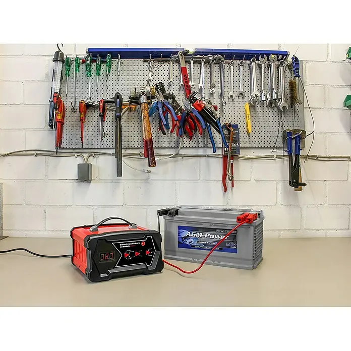 APA Cargador para el taller - Corriente de carga: 2 A - 12 A - Un cargador de batería rojo está conectado a una batería de coche gris. Las herramientas cuelgan de un soporte de pared al fondo.