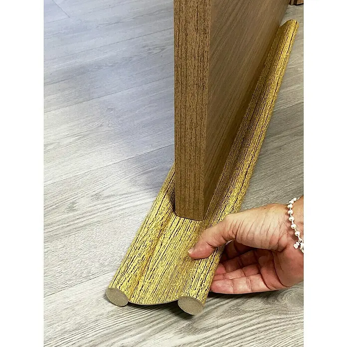 Junta para puerta Rodillo - Roble, Largo: 95 cm - Esta imagen muestra una pieza de moldura de madera en la base de un mueble, fijada al suelo, siendo tocada por una mano.