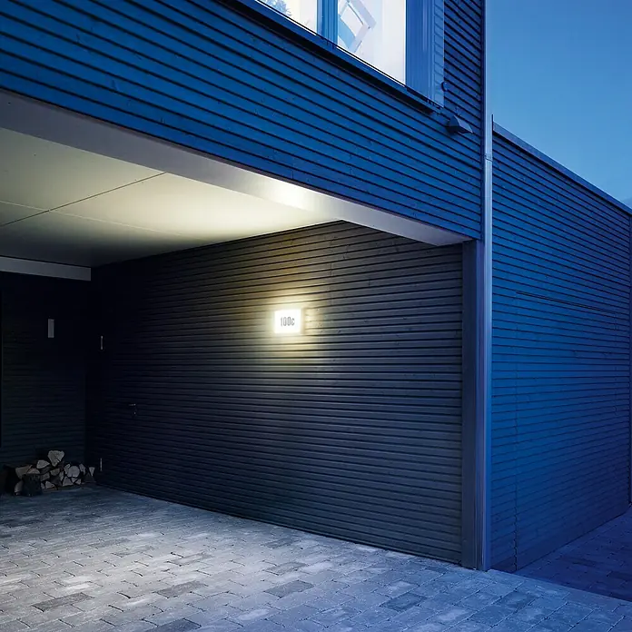 Steinel LED-Außenwandleuchte L 1 N - 4,9 W, 300 x 195 x 53 mm, Weiß, IP54 - Eine moderne Hausnummernleuchte beleuchtet eine dunkle Holzfassade. Das Licht ist hell und einladend.