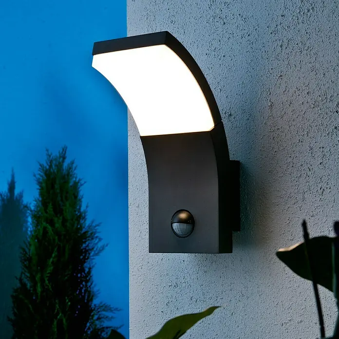 Starlux LED-Sensor-Außenwandleuchte Vincenza - 7 W, L x B x H: 122 x 84 x 190 mm, Silbergrau, Opal, Warmweiß - Nahaufnahme einer modernen schwarzen Außenwandleuchte mit Bewegungssensor, die an einer grauen Wand montiert ist und helles Licht ausstrahlt.