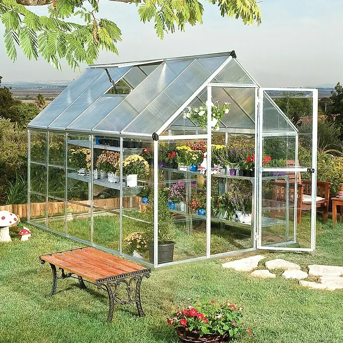 Palram – Canopia Gewächshaus Hybrid - Außenmaß inkl. Dachüberstand (B x T): 185 x 306 cm, 0,7 mm, Silber - Ein Gewächshaus im Garten, gefüllt mit bunten Blumen auf Regalen. Davor eine Bank und ein Blumenkorb.