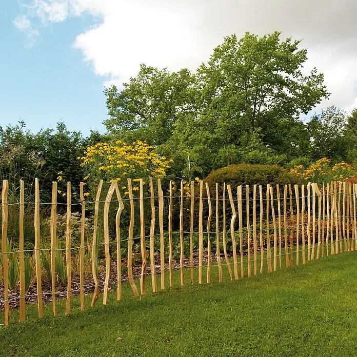 Kiehnholz Staketenzaun - 500 x 90 cm, Haselnuss - Ein Holzzaun aus naturbelassenen Stämmen umzäunt einen Garten mit üppiger Bepflanzung, der Schutz und rustikalen Charme bietet.