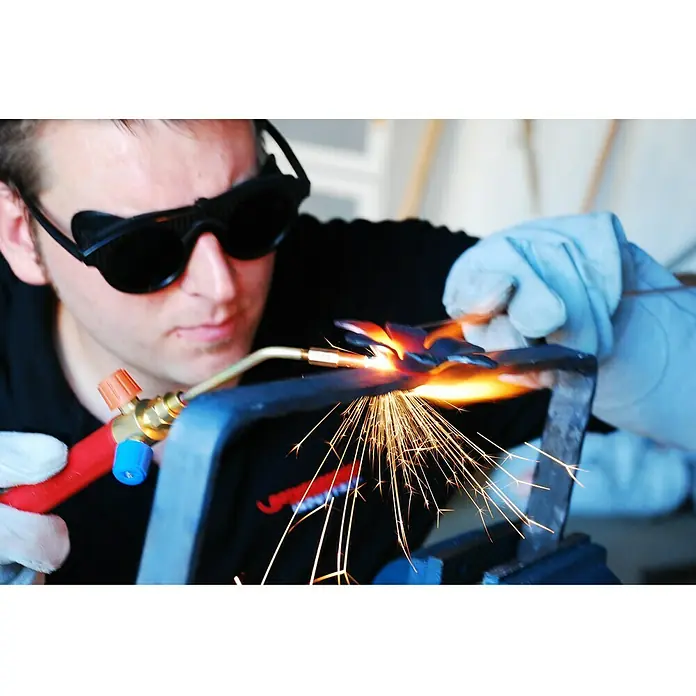 Rothenberger Industrial Equipo de soldadura autógena ROXY KIT Plus - 1.250 °C - Imagen de un hombre con gafas de protección y guantes, trabajando una pieza de metal con un soplete, saltando chispas.