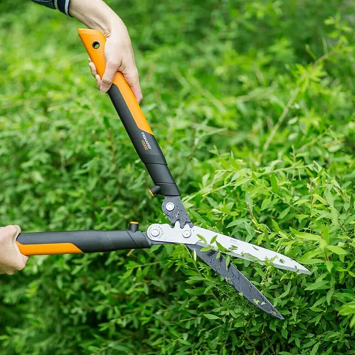 Fiskars Getriebe-Heckenschere Powergear X HSX 92 - Länge: 63 cm - Eine Person schneidet mit einer Fiskars Heckenschere einen Busch im Garten. Die Schere hat orangefarbene Griffe und silberne Klingen.
