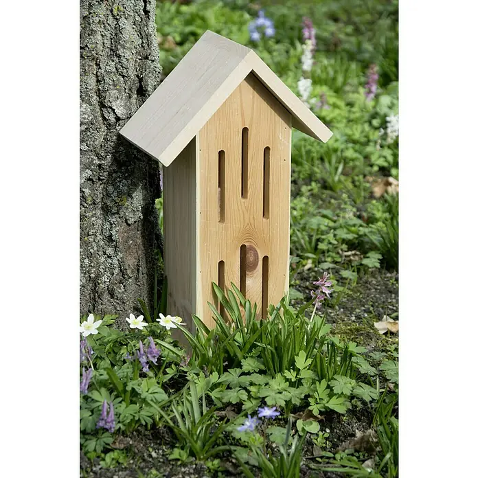 Dobar Schmetterlingsunterschlupf Sonnenfalter - 23 x 14 x 38 cm, Kiefer - Ein hölzernes Insektenhotel steht am Fuße eines Baumes, umgeben von grünen Pflanzen und kleinen Blüten. Ideal für den naturnahen Garten.