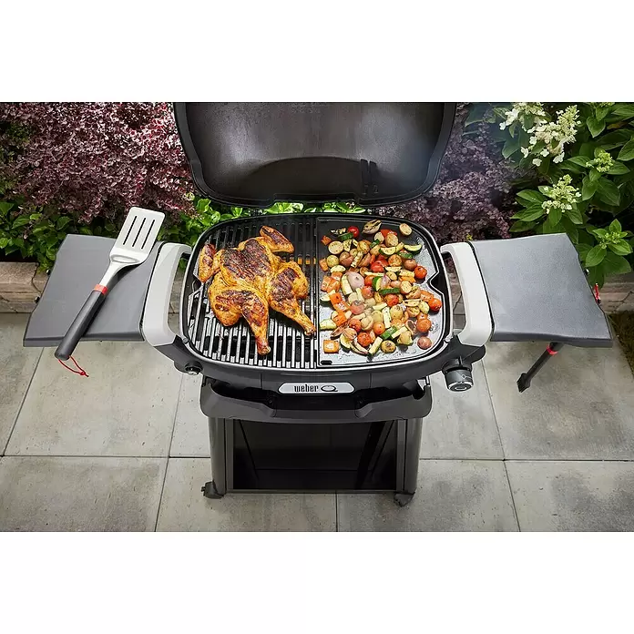 Weber Q Grillplatte Plancha - Passend für: Weber Q 2100N, Q 2200N, Q 2800N+, Gusseisen - Ein grauer Weber Q Grill im Freien, auf dessen Rost ein Hähnchen und gemischtes Gemüse garen. Ein Spatel liegt auf dem linken Seitentisch.