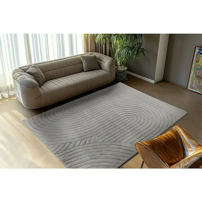 Fellteppich Zen - Silber, 170 x 120 cm, 100 % Polyester - Ein grauer Teppich mit wellenförmigem Muster liegt vor einem Sofa, bringt Eleganz in den Raum und sorgt für ein modernes Ambiente.