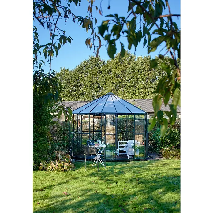 Halls Gewächshaus Atrium - Außenmaß inkl. Dachüberstand (B x T): 380 x 380 cm, Material Seitenwand: Glas, 3 mm, Grün - Ein elegantes Gewächshaus im Garten, umgeben von Grün, mit einem Tisch und Stühlen für entspannte Momente. Das Gewächshaus bietet Schutz und Stil.