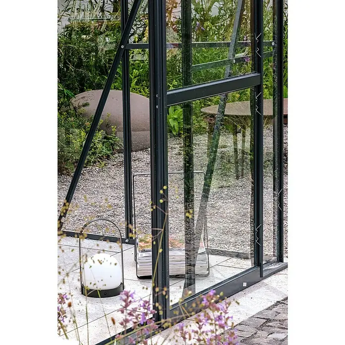 Halls Gewächshaus Magnum 108 - Außenmaß inkl. Dachüberstand (B x T): 257 x 322 cm, 3 mm, Grün, Sicherheitsglas - Detailaufnahme eines Gewächshauses mit Glaswänden und schwarzem Rahmen, ergänzt durch dekorative Elemente wie eine Lampe und Zeitschriften.