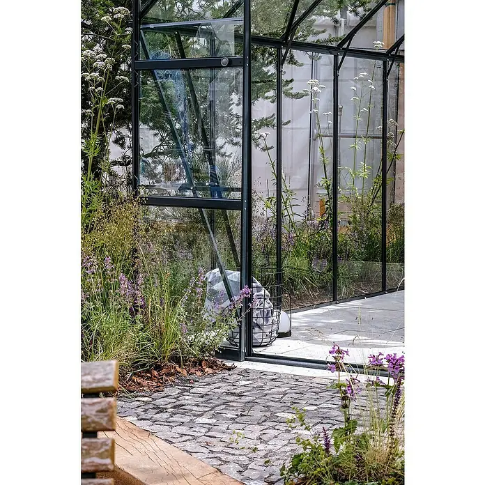 Halls Gewächshaus Magnum 108 - Außenmaß inkl. Dachüberstand (B x T): 257 x 322 cm, 3 mm, Grün, Sicherheitsglas - Eingang zu einem modernen Gewächshaus mit Glaswänden und Metallrahmen, umgeben von üppiger Vegetation und einem gepflasterten Weg.