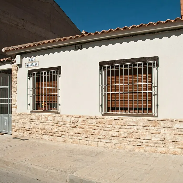 Revestimiento de piedra Silleria Cobre - Cobre, Cantidad total: 4 pzs. - Vista exterior de una casa con paredes blancas, dos ventanas con rejas y un letrero que dice "Casa Santiago".