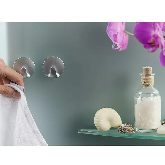 Tesa Powerstrips Gancho autoadhesivo Mini Redondo - Plateado - Imagen de dos ganchos de color plateado en una pared, con una mano colgando una toalla blanca y artículos de baño en una estantería de cristal.