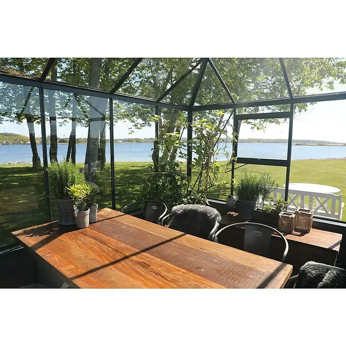 Juliana Gewächshaus Grand Oase - Außenmaß inkl. Dachüberstand (B x T): 368 x 511 cm, 3 mm, Anthrazit/Schwarz, Für Mauerfundament - Einladende Inneneinrichtung eines Gewächshauses mit Holztisch, Stühlen mit Fellauflagen und Blick auf den See, Pflanzen in Töpfen verschönern den Raum.