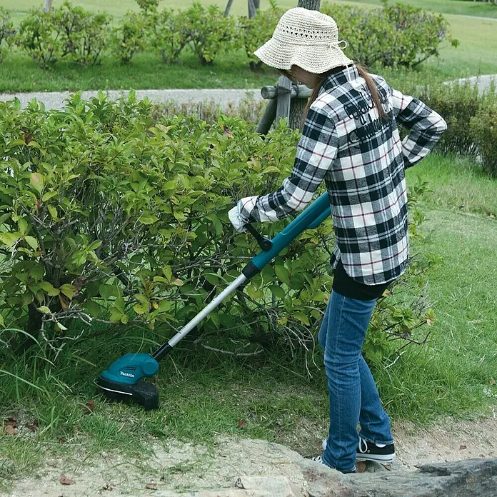 Makita LXT 18V Akku-Rasentrimmer DUR181Z - 18 V, Li-Ionen, Ohne Akku, Schnittbreite Faden: 260 mm - Eine Frau schneidet mit einer Makita Akku-Grasschere im Garten Büsche. Sie trägt eine karierte Jacke und einen Sonnenhut.
