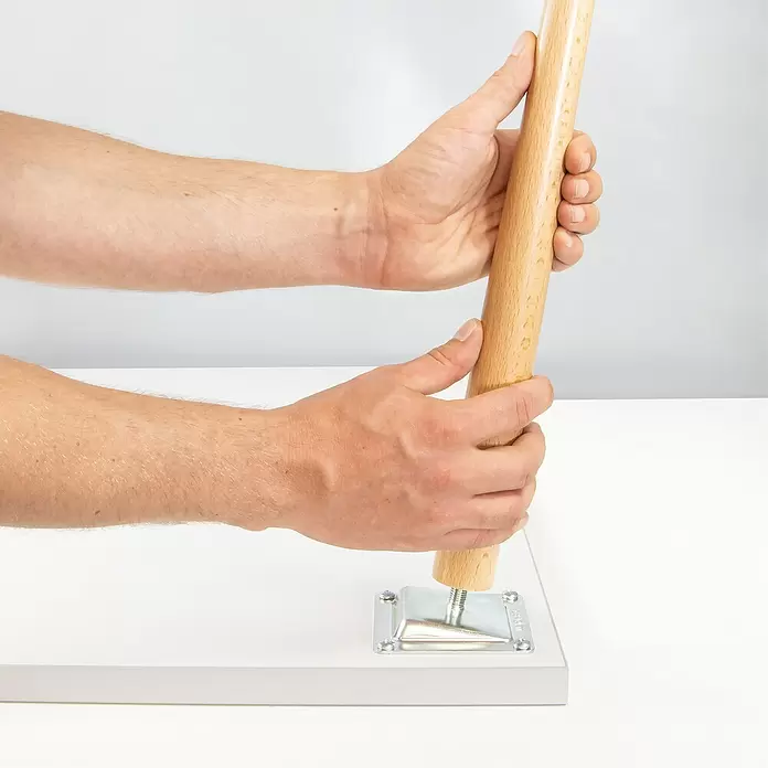 Wagner System Möbelfuß - Ø x H: 35 x 210 mm, Holz - Hände befestigen einen hellbraunen Holzmöbelfuß mit Metallbefestigungsplatte an einer weißen Platte.