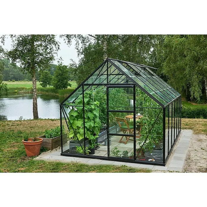 Halls Gewächshaus Universal 128 - Inkl. Fundamentrahmen - Ein Gewächshaus mit Pflanzen, Gartentisch und Stühlen steht in einem Garten. Das Gewächshaus ist aus Glas und Metall gefertigt.