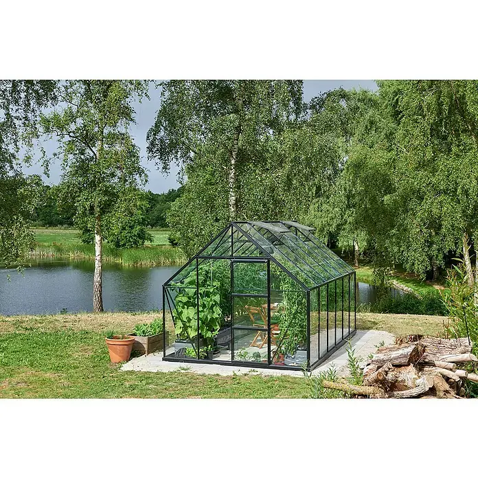 Halls Gewächshaus Universal 128 - Inkl. Fundamentrahmen - Ein Gewächshaus mit Pflanzen steht im Garten neben einem Teich, umgeben von Bäumen und Grünflächen.