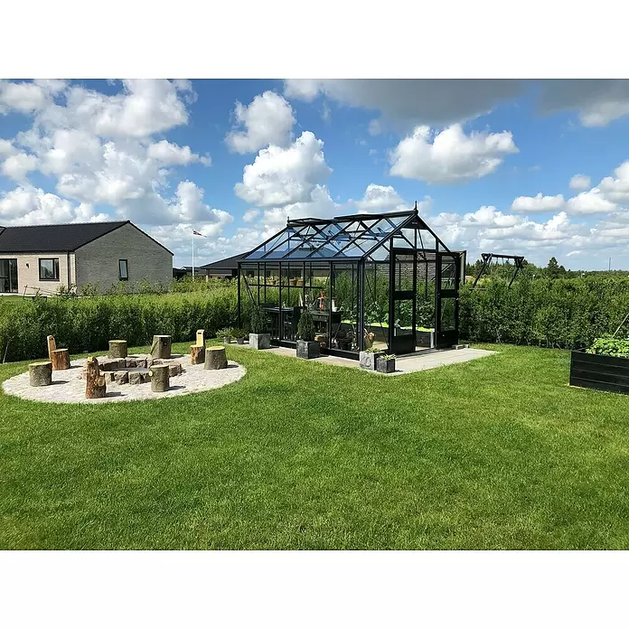 Juliana Gewächshaus Premium - Außenmaß inkl. Dachüberstand (B x T): 296 x 368 cm, 3 mm, Anthrazit, Einscheibensicherheitsglas - Schwarz gerahmtes Glasgewächshaus in grünem Garten, innen mit Pflanzen und Tisch. In der Nähe ist eine Feuerstelle mit Holzstämmen.