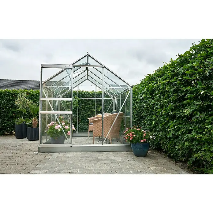 Halls Gewächshaus Popular 86 - Außenmaß inkl. Dachüberstand (B x T): 193 x 257 cm, Material Seitenwand: Glas, 3 mm, Silber - Ein Gewächshaus aus Glas und Metall mit Pflanzen und einem Stuhl darin, umgeben von Grün.