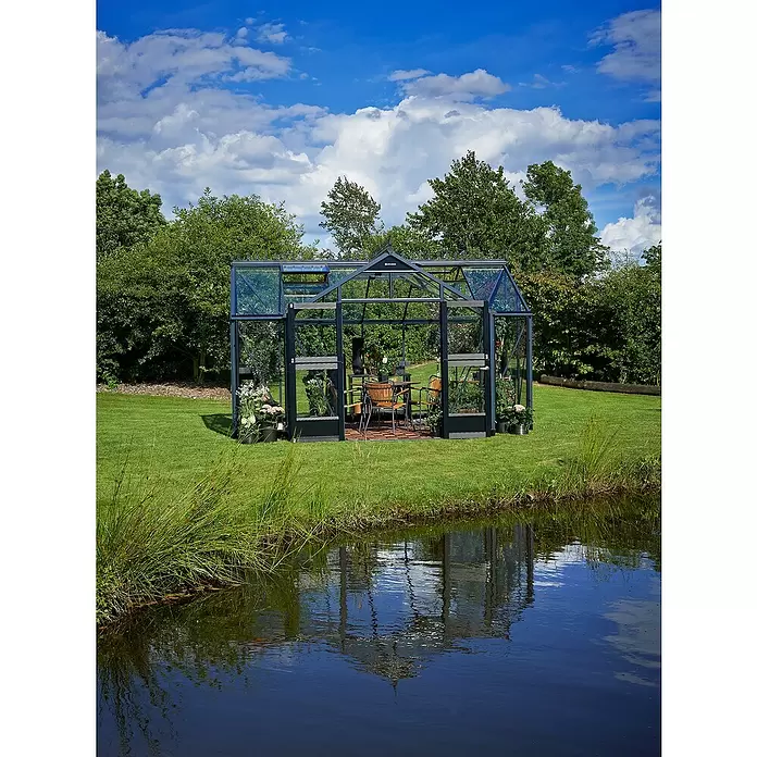 Juliana Gewächshaus Orangerie - Außenmaß inkl. Dachüberstand (B x T): 439 x 296 cm, Material Seitenwand: Glas, 3 mm, Anthrazit - Ein dunkel gerahmtes Glasgewächshaus mit Sitzbereich, auf grünem Rasen neben einem Teich stehend.