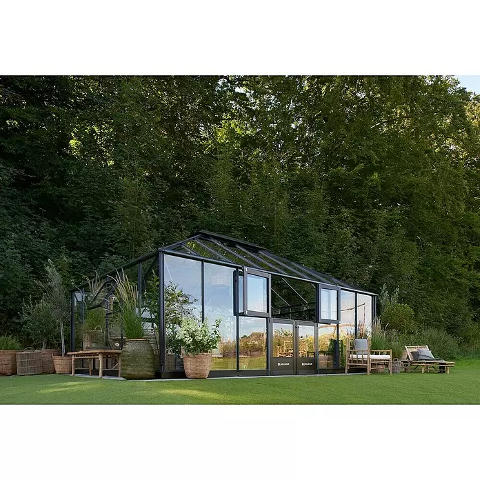Juliana Gewächshaus Grand Oase - Material Seitenwand: Glas, 3 mm, Anthrazit - Ein Glashaus mit schwarzem Rahmen steht auf grünem Rasen in einem Garten, umgeben von Bäumen und Gartenmöbeln.
