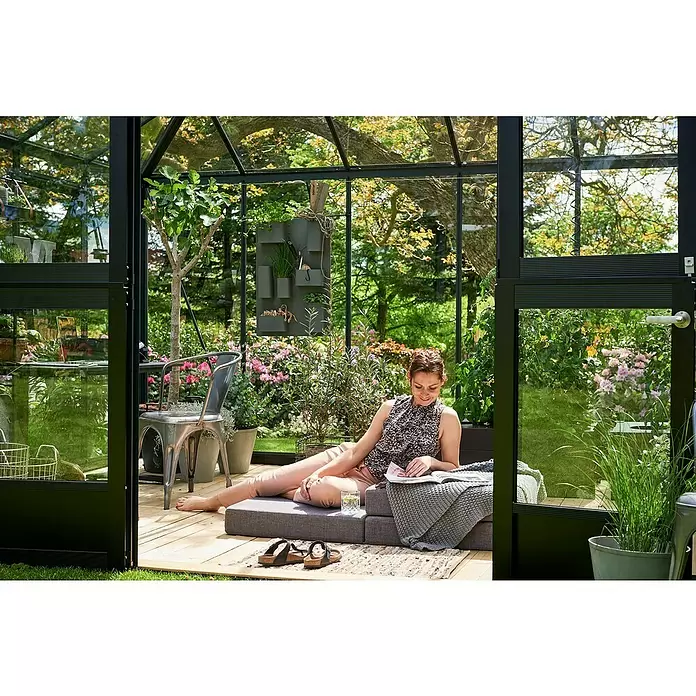 Juliana Gewächshaus Grand Oase - Außenmaß inkl. Dachüberstand (B x T): 439 x 296 cm - Frau entspannt in einem modernen Gewächshaus mit schwarzem Rahmen, Glaswänden und Holzboden. Sie liest ein Buch auf grauen Polstern.