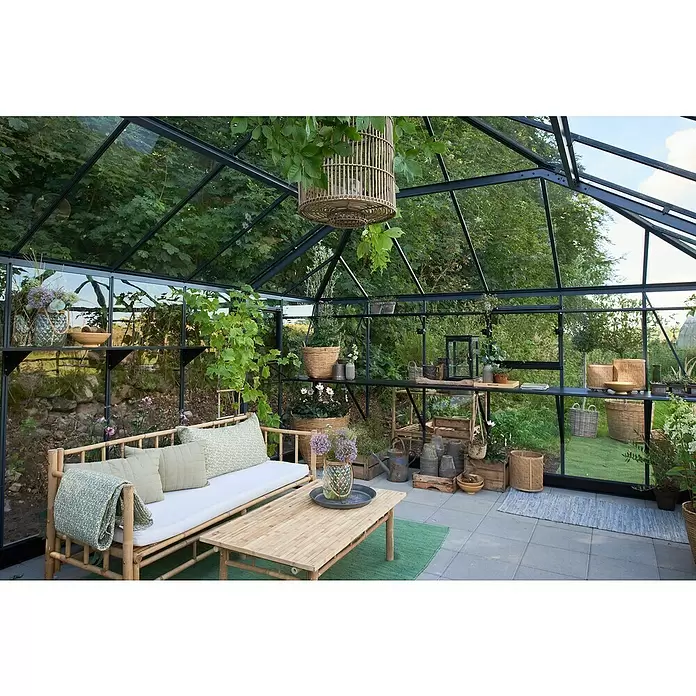 Juliana Gewächshaus Grand Oase - Material Seitenwand: Glas, 3 mm, Anthrazit - Geräumiges Gewächshaus mit schwarzem Rahmen und Glaspaneelen, ausgestattet mit Bambus-Lounge-Möbeln und Gartenutensilien für einen gemütlichen Wohnbereich im Freien.