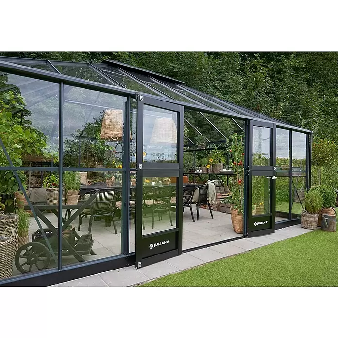 Juliana Gewächshaus Grand Oase - Material Seitenwand: Glas, 3 mm, Anthrazit - Großes, modernes Gewächshaus mit dunklem Rahmen, Glaspaneelen, Pflanzen und Essbereich im Garten.