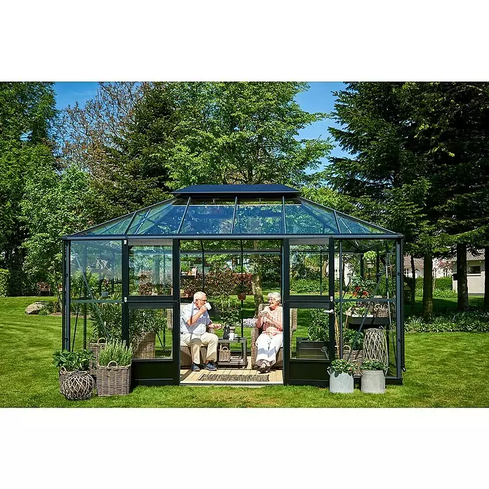 Juliana Gewächshaus Grand Oase - Außenmaß inkl. Dachüberstand (B x T): 439 x 296 cm - Dunkelgerahmtes Glasgewächshaus in einem Garten, in dem zwei Personen Kaffee trinken, umgeben von Pflanzen.