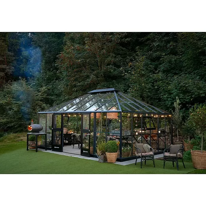 Juliana Gewächshaus Grand Oase - Material Seitenwand: Glas, 3 mm, Anthrazit - Ein schwarz gerahmtes Gewächshaus aus Glas, beleuchtet in einem üppigen Garten bei Dämmerung, mit einem rauchenden Ofen daneben.