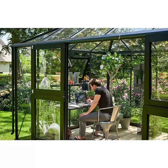 Juliana Gewächshaus Grand Oase - Außenmaß inkl. Dachüberstand (B x T): 439 x 296 cm - Schwarz gerahmtes Glasgewächshaus, als Arbeitsbereich mit Mann und Pflanzen genutzt.