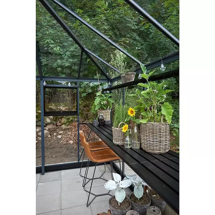 Juliana Gewächshaus Grand Oase - Material Seitenwand: Glas, 3 mm, Anthrazit - Modernes schwarzes Gewächshaus mit Glaspaneelen, schwarzem Tisch und braunen Stühlen, für Pflanzen und Entspannung im Garten.