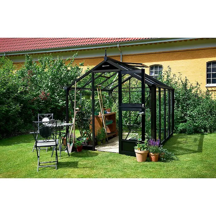 Juliana Gewächshaus Compact - Außenmaß inkl. Dachüberstand (B x T): 224 x 296 cm, Material Seitenwand: Glas, 3 mm, Schwarz - Ein elegantes Juliana Gewächshaus aus schwarzem Rahmen und Glas steht in einem üppigen Garten, ideal für Hobbygärtner und Pflanzenliebhaber.
