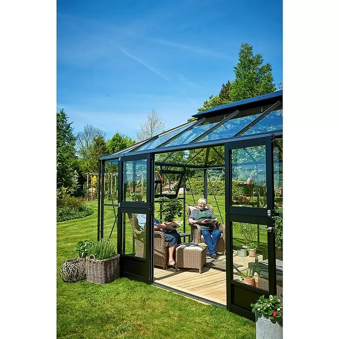 Juliana Gewächshaus Grand Oase - Außenmaß inkl. Dachüberstand (B x T): 439 x 296 cm - Schwarz gerahmtes Glasgewächshaus mit offenen Türen, in dem zwei Personen auf Korbstühlen in einem sonnigen Garten entspannen.